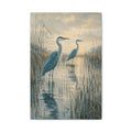 Two in the Reeds canvas print — Two herons stand among reeds and still water, one slightly forward and one farther back.